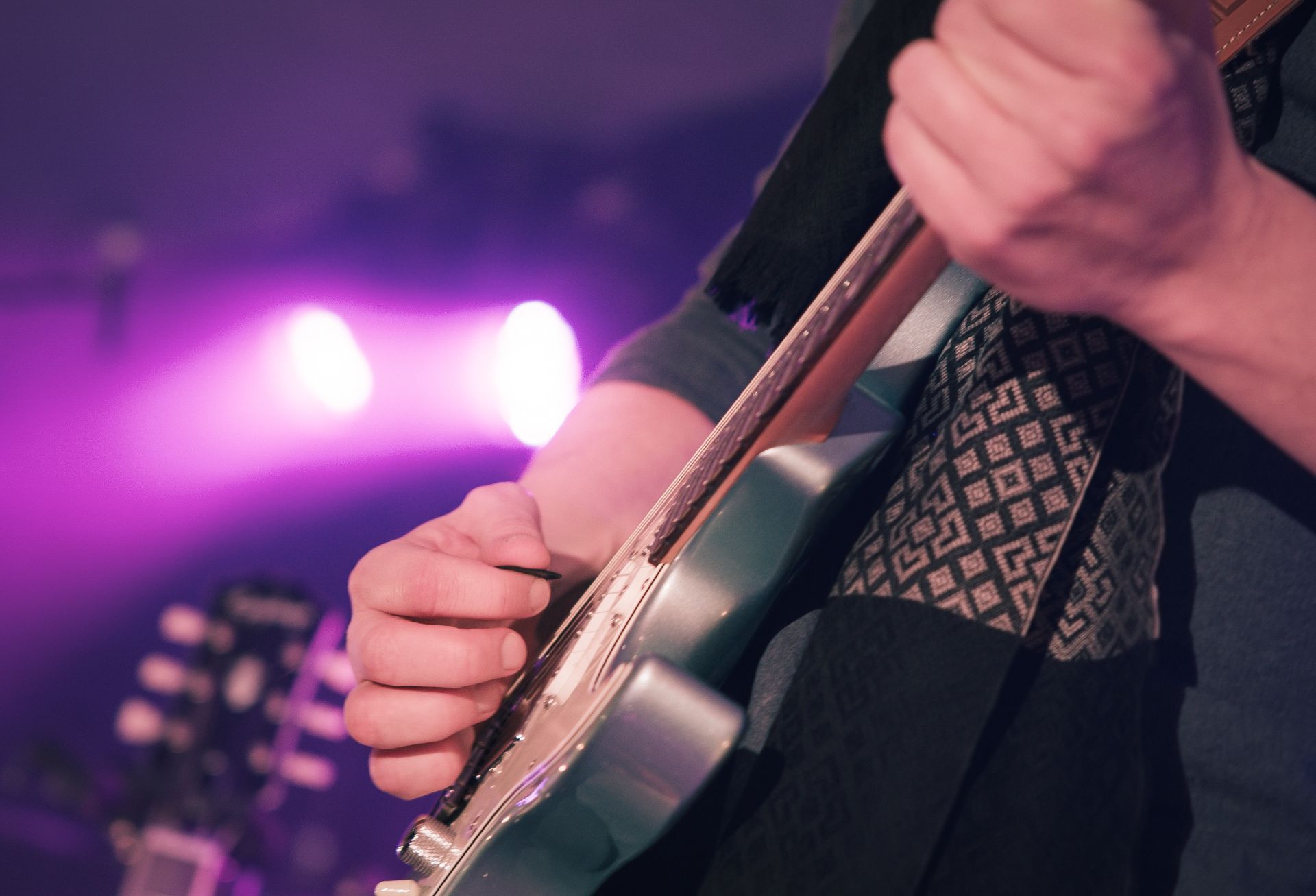 En mann spiller en elektrisk gitar på en scene med lilla lys bak seg.