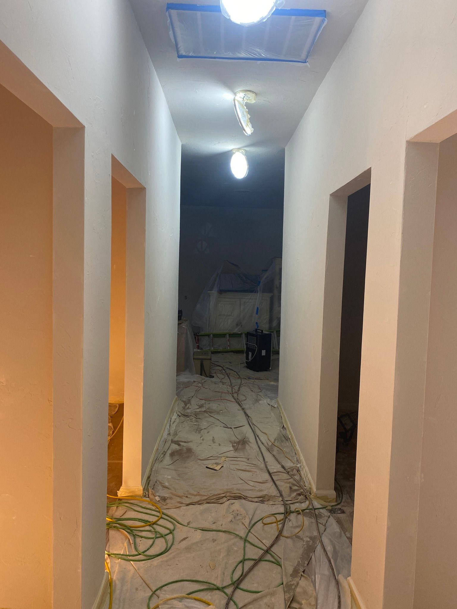 Narrow hallway under renovation with exposed wires, unfinished walls, and work lights hanging overhead
