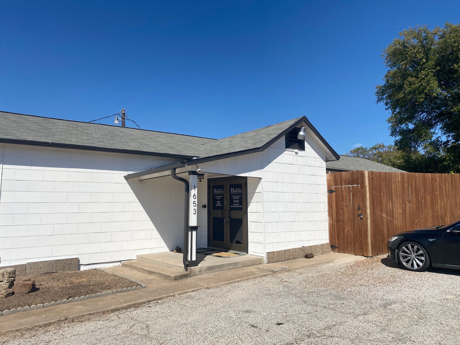 White building entrance with small awning, wooden fence, gravel driveway, and a black car under blue sky