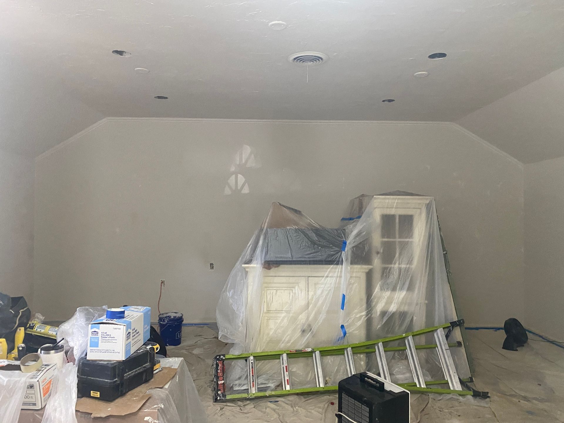 Garage-like room with plastic-covered furniture, a ladder, and construction materials spread across the floor.