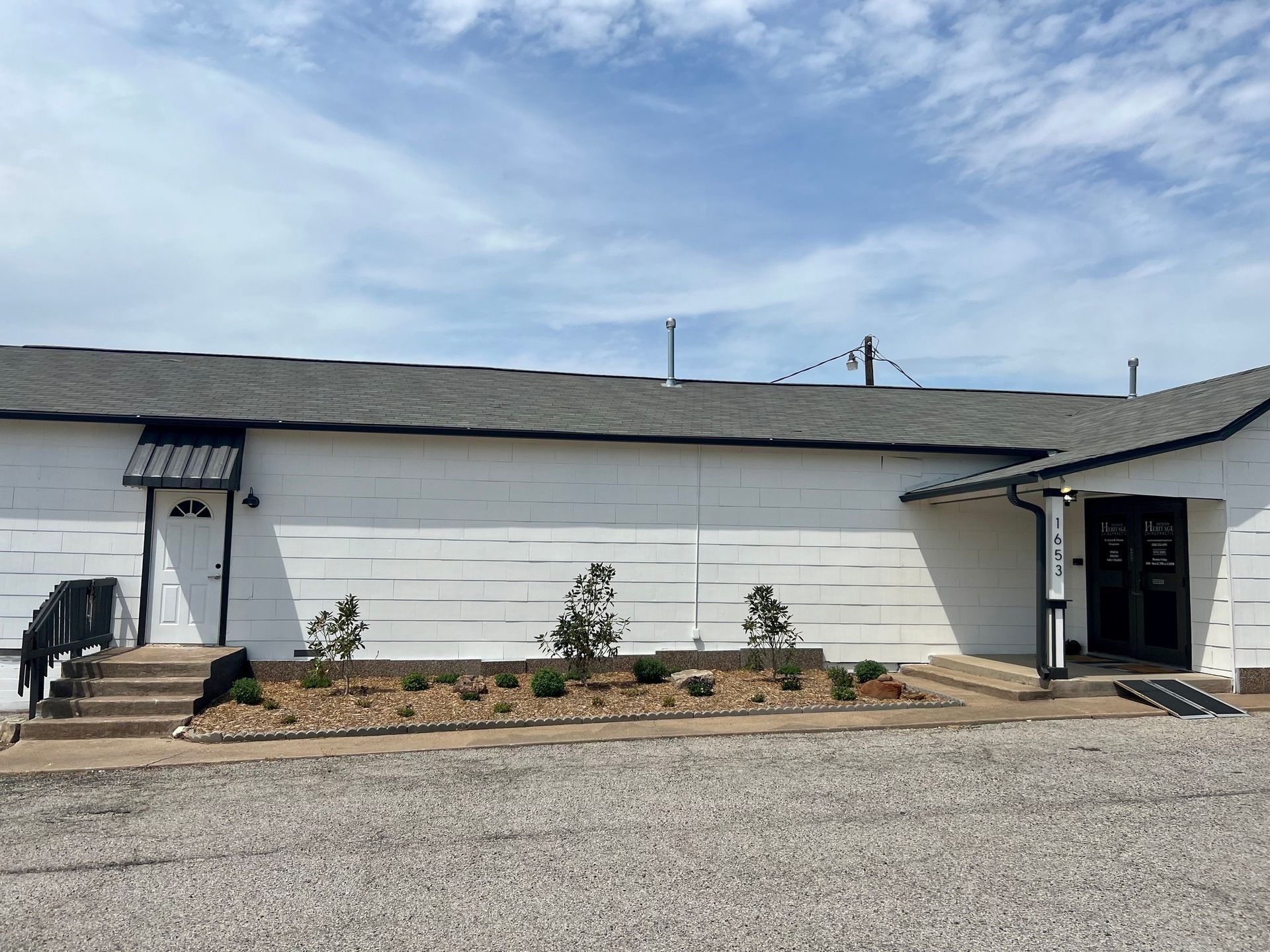 White industrial building with a black roof, small front entry, and sparse landscaping under a blue sky