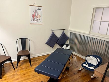 Small room with blue massage table, two chairs, wall art, and a baby bouncer near a window.