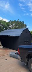 Black shed with a gray roof in a driveway under a blue sky, with a pickup truck in the foreground