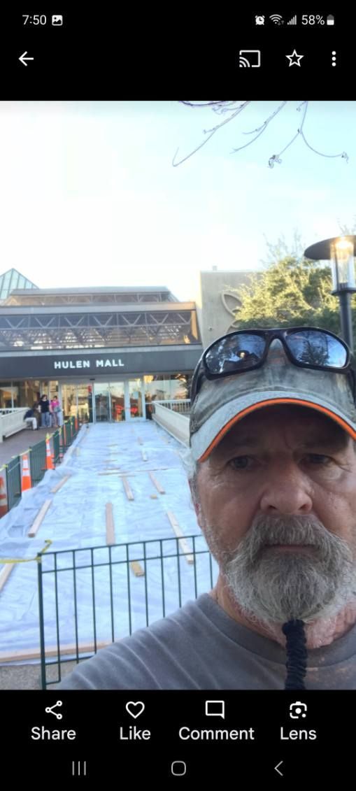 Man in cap and sunglasses taking a selfie in front of a ticketed entrance and metal barricades