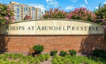 Stone sign reading “Shops at Arundel Preserve” with flowers, shrubs, and apartment buildings behind it