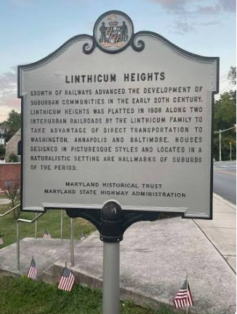 Historic Linthicum Heights roadside marker with black text on a gray sign beside a sidewalk and small flags