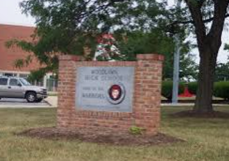 Brick school entrance sign beside a tree and building, with a white truck parked nearby.