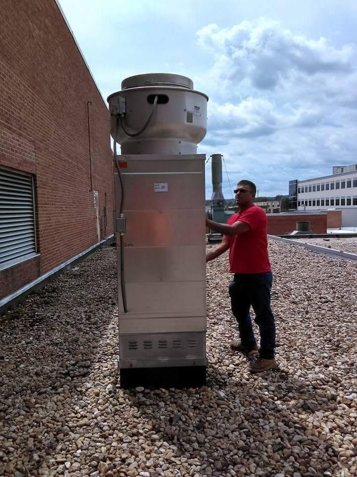 Man in red shirt beside large silver HVAC unit on a gravel rooftop near a brick wall