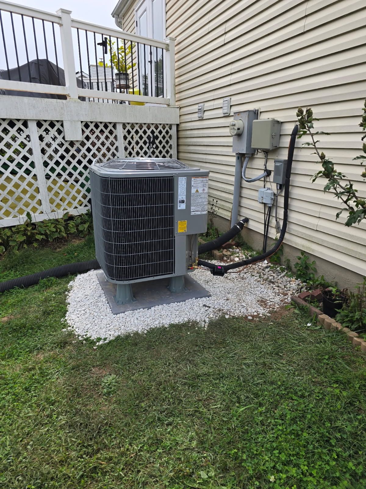 Outdoor air conditioner unit beside beige house wall on gravel pad, with grass and lattice fence nearby Outdoor air conditioner unit beside beige house wall on gravel pad, with grass and lattice fence nearby