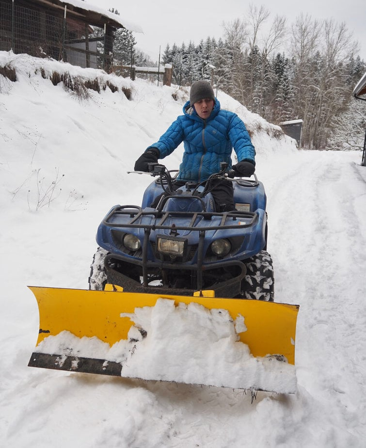 Person on blue ATV with yellow plow clearing snow on a driveway.