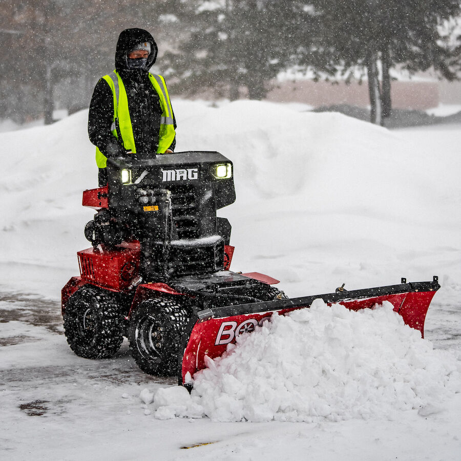 Person operating a red snowplow, clearing snow from a driveway, wearing a reflective vest in winter.