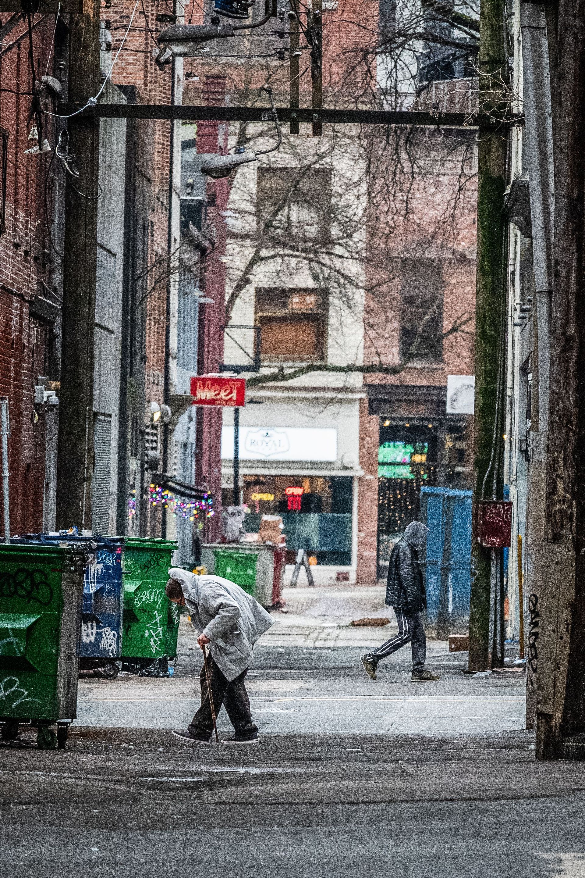 Snowy urban alley with a person sweeping near green bins and brick buildings in the background