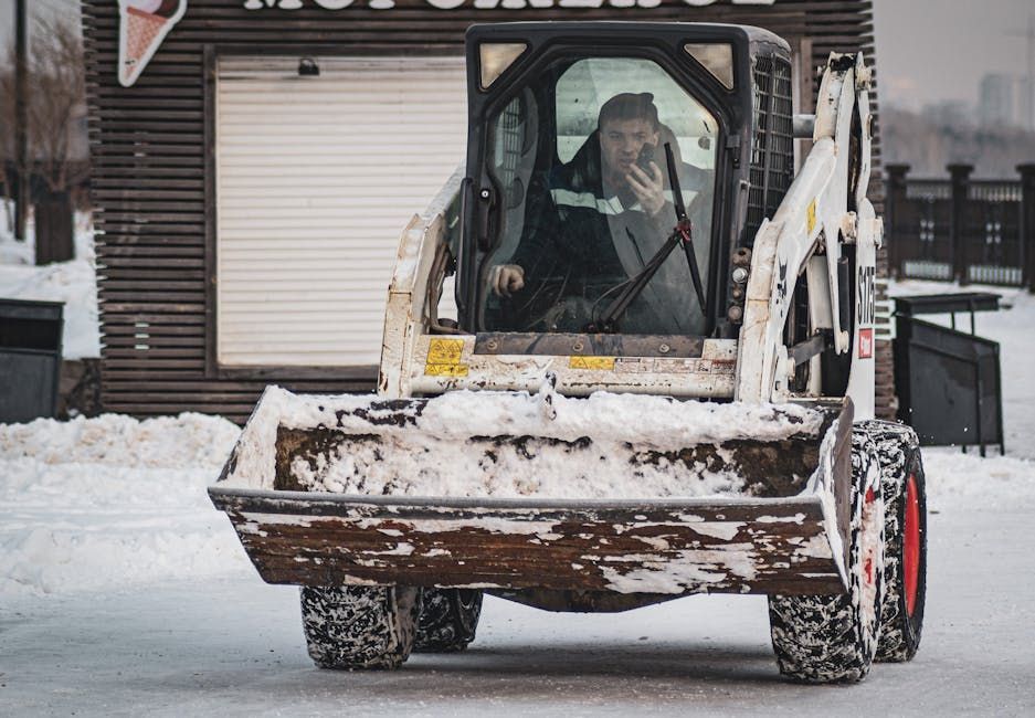 A man operates a Bobcat skid-steer loader, scooping snow. The vehicle is white, set in a snowy outdoor area.