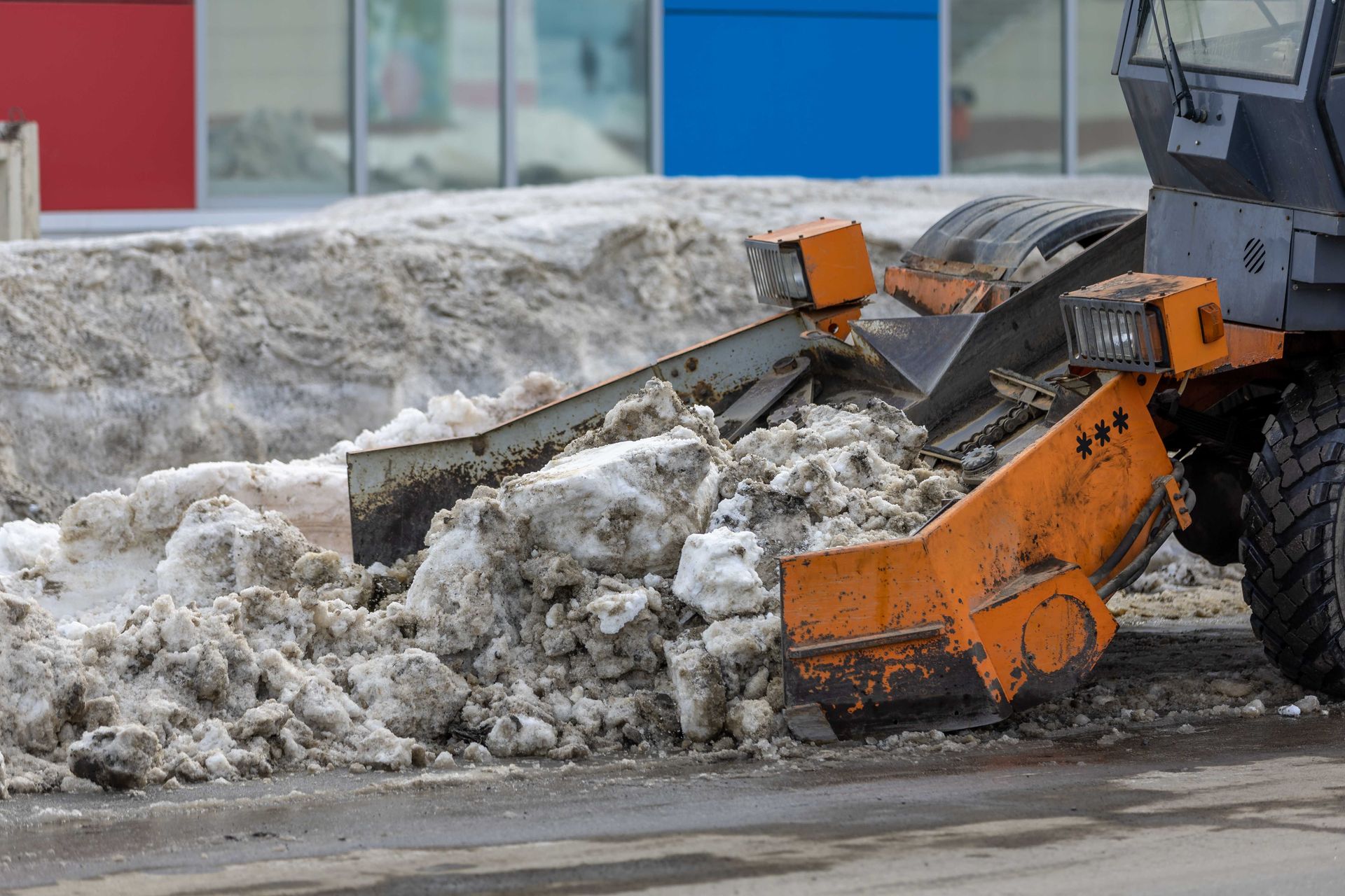 Orange snowplow clearing a dirty snowbank along a roadside near colorful buildings