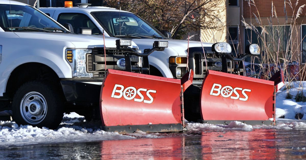 Two white pickup trucks with red BOSS snowplows clearing icy pavement outdoors.