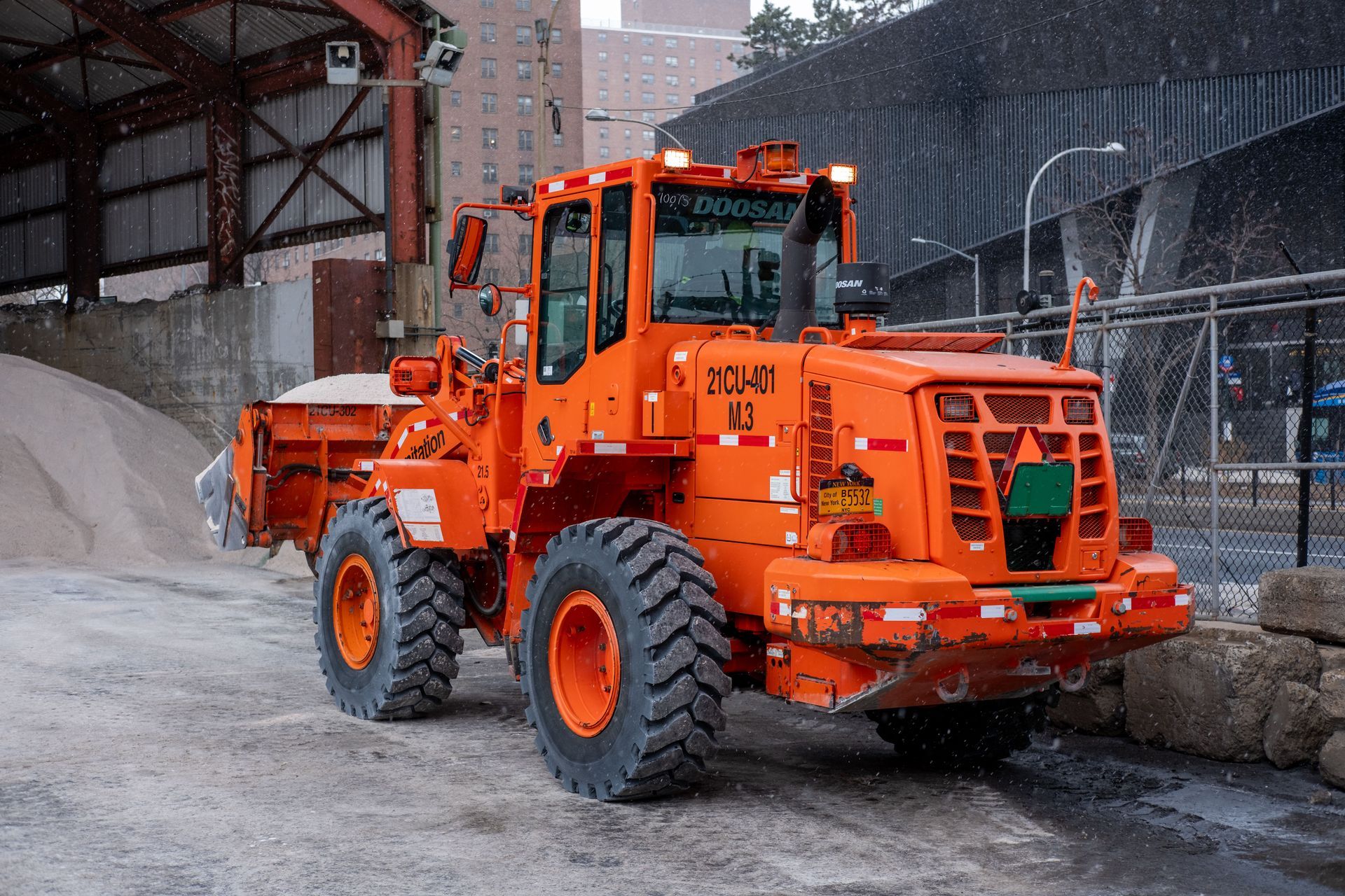 Bright orange construction loader carrying snow beside concrete blocks in an industrial yard