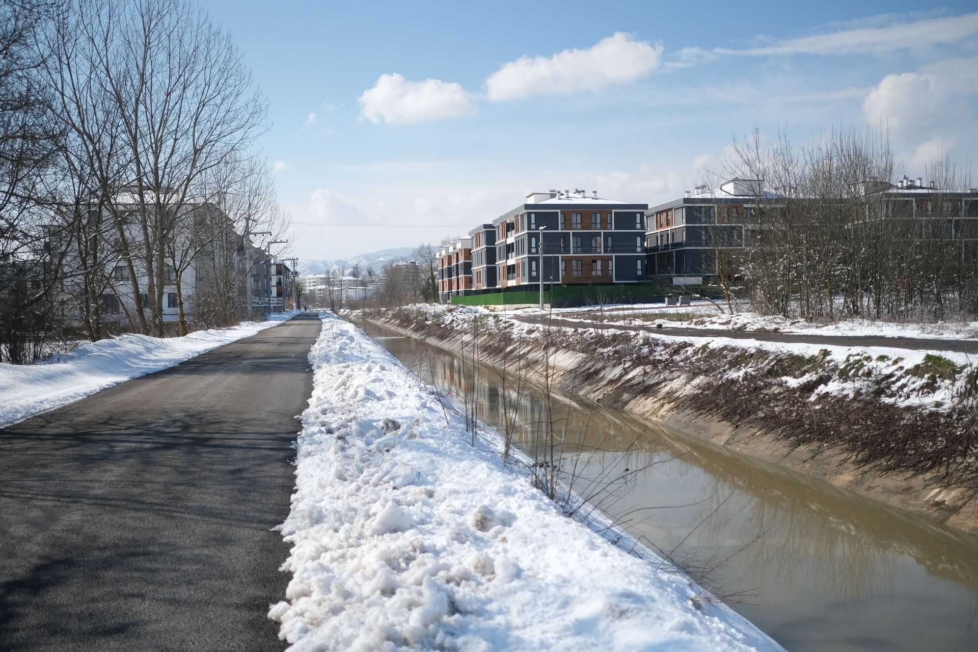 Snowy canal path beside a frozen waterway and modern buildings under a blue sky