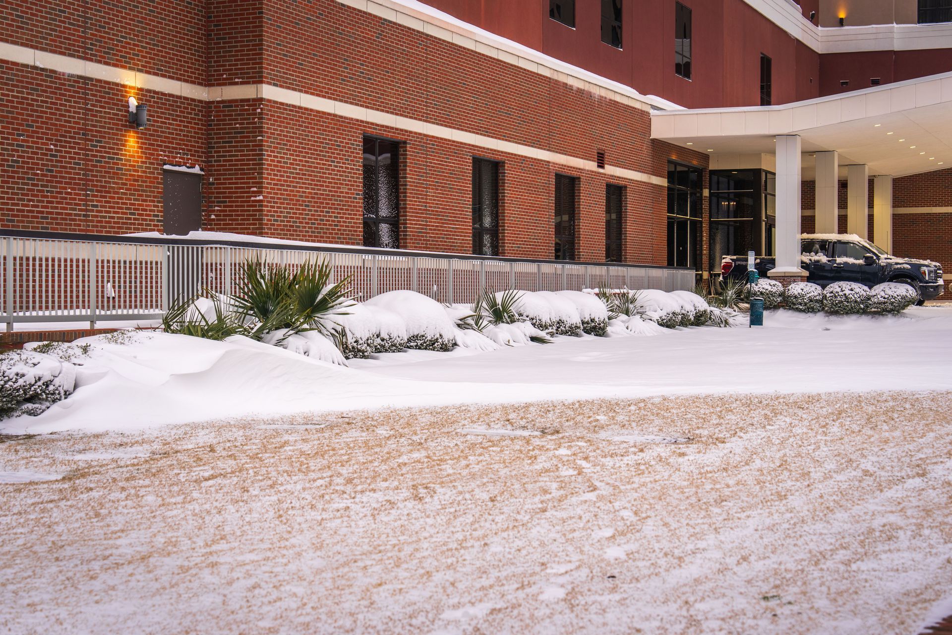 Snow-covered street beside a brick building with parked cars and a lit entrance