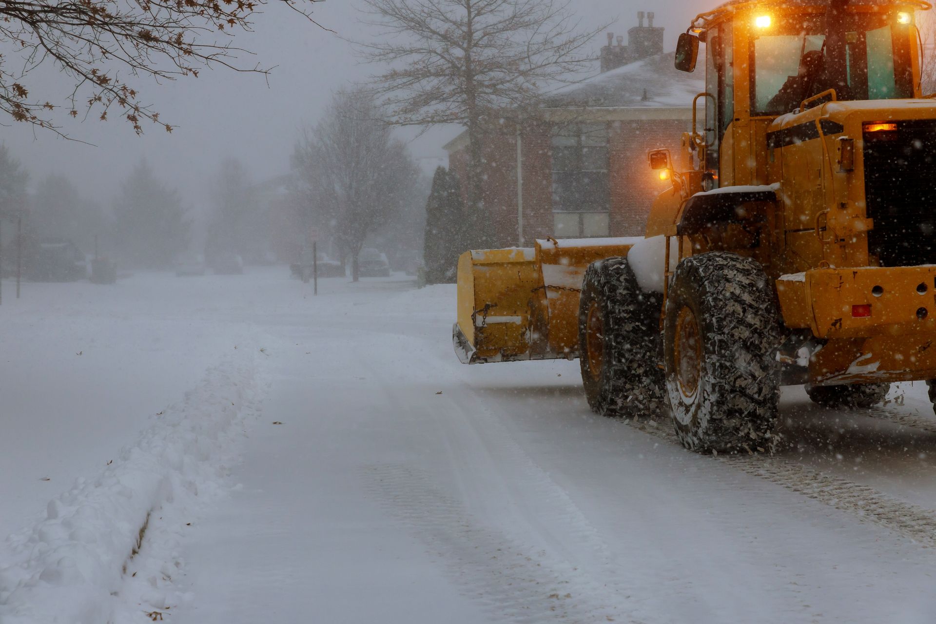 Person operating a red snowplow, clearing snow from a driveway, wearing a reflective vest in winter.