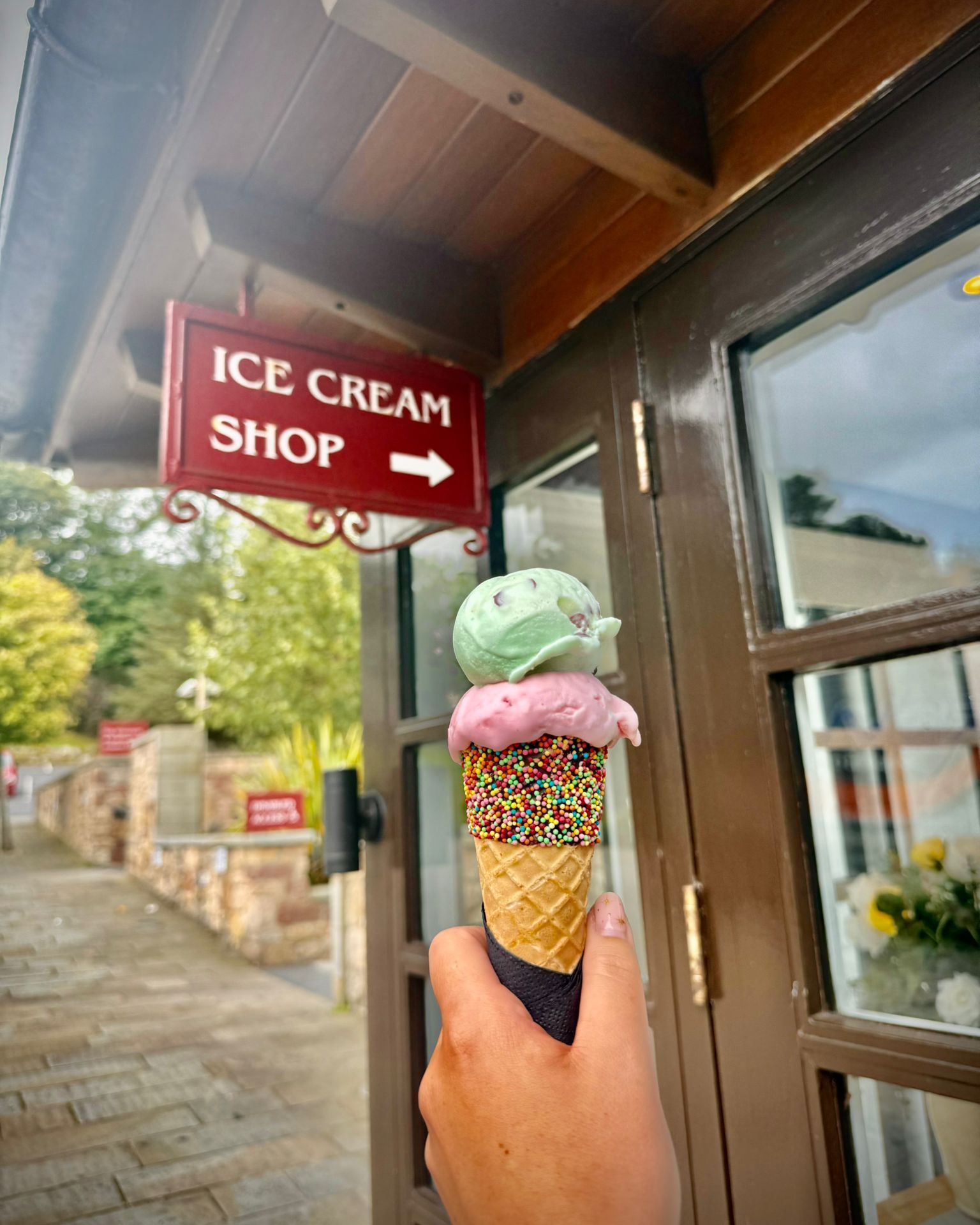 an image of a hand holding a waffle cone with pink and green ice cream outside an ice cream shop sign