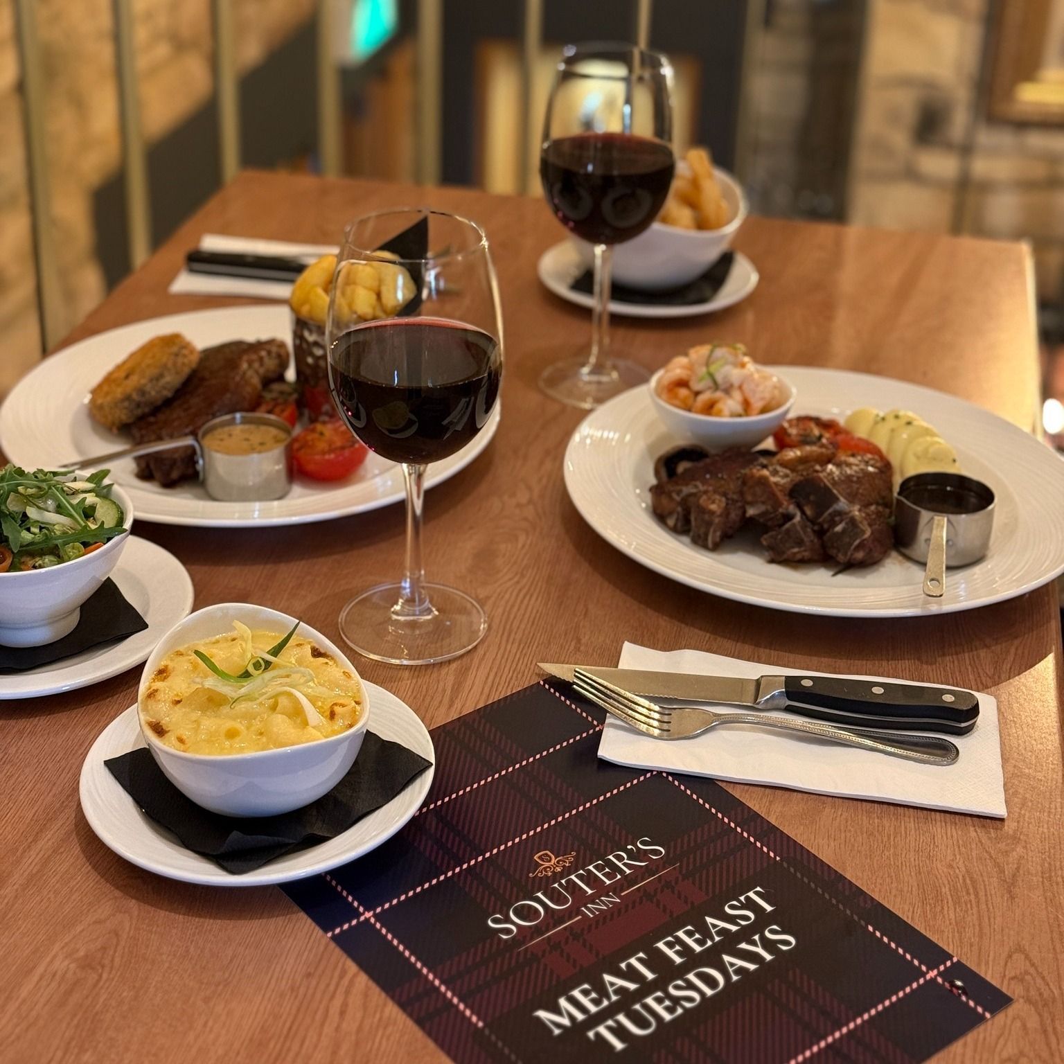 an image showing a restaurant table with steak dinner, macaroni cheese side, red wine, and a menu for a meat feast special