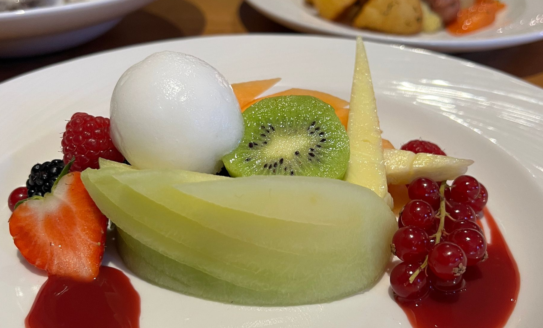 an image of a dessert plate with melon, kiwi, strawberries, raspberries, and a scoop of sorbet in red sauce