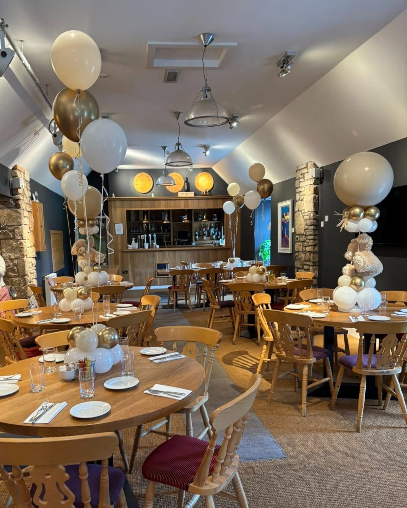 Decorated restaurant dining room with round tables, white and gold balloons, and a bar at the back