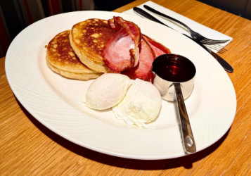 a close up of a plate of breakfast with pancakes, bacon, poached eggs and a metal pouring container of syrup on the side.