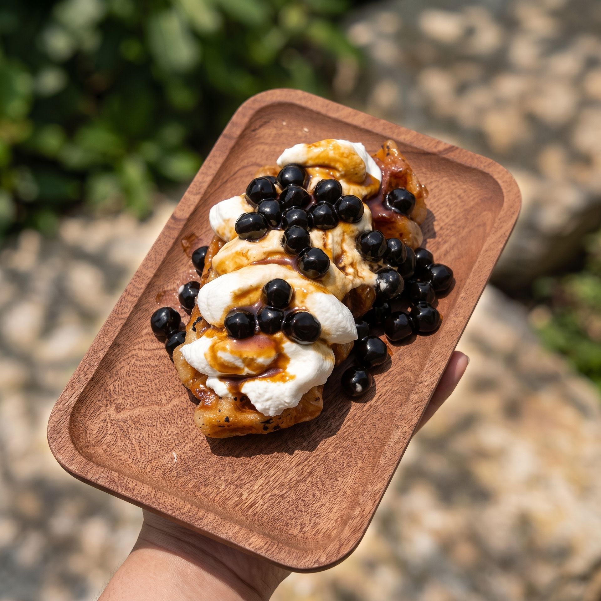 A dessert on a wooden tray topped with whipped cream, caramel sauce, and black berries outdoors.