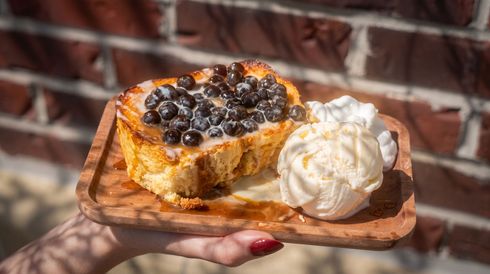 Hand holding a wooden tray with berry-topped cake and a scoop of vanilla ice cream against a brick wall