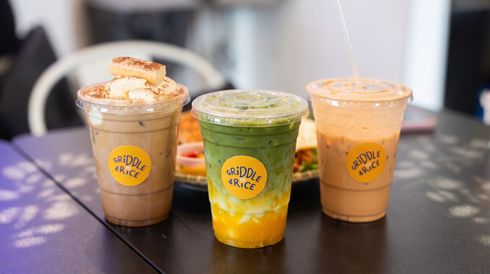 Three iced drinks in clear cups with yellow logos on a table, including brown, green, and peach-colored drinks.