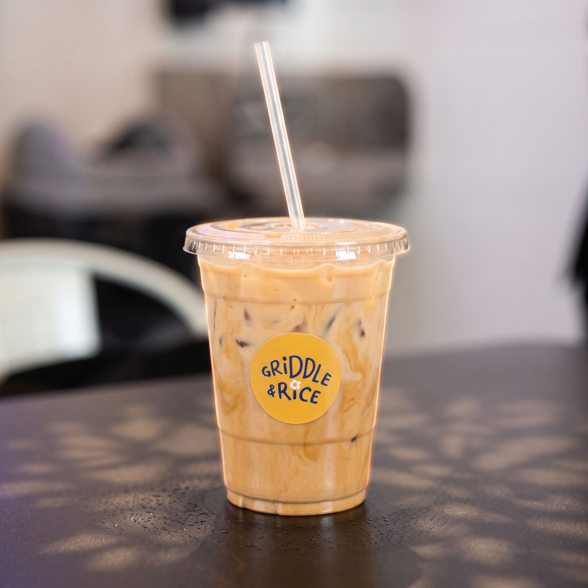 Iced coffee in a clear plastic cup with a straw on a tabletop, labeled “Griddle & Rice.”