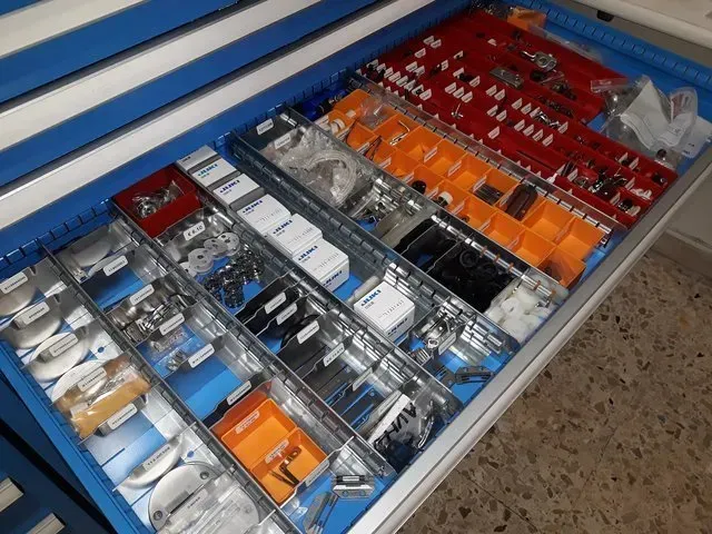Organizzatore a cassetto aperto con vari piccoli attrezzi e accessori in scomparti rossi, arancioni, blu e bianchi.