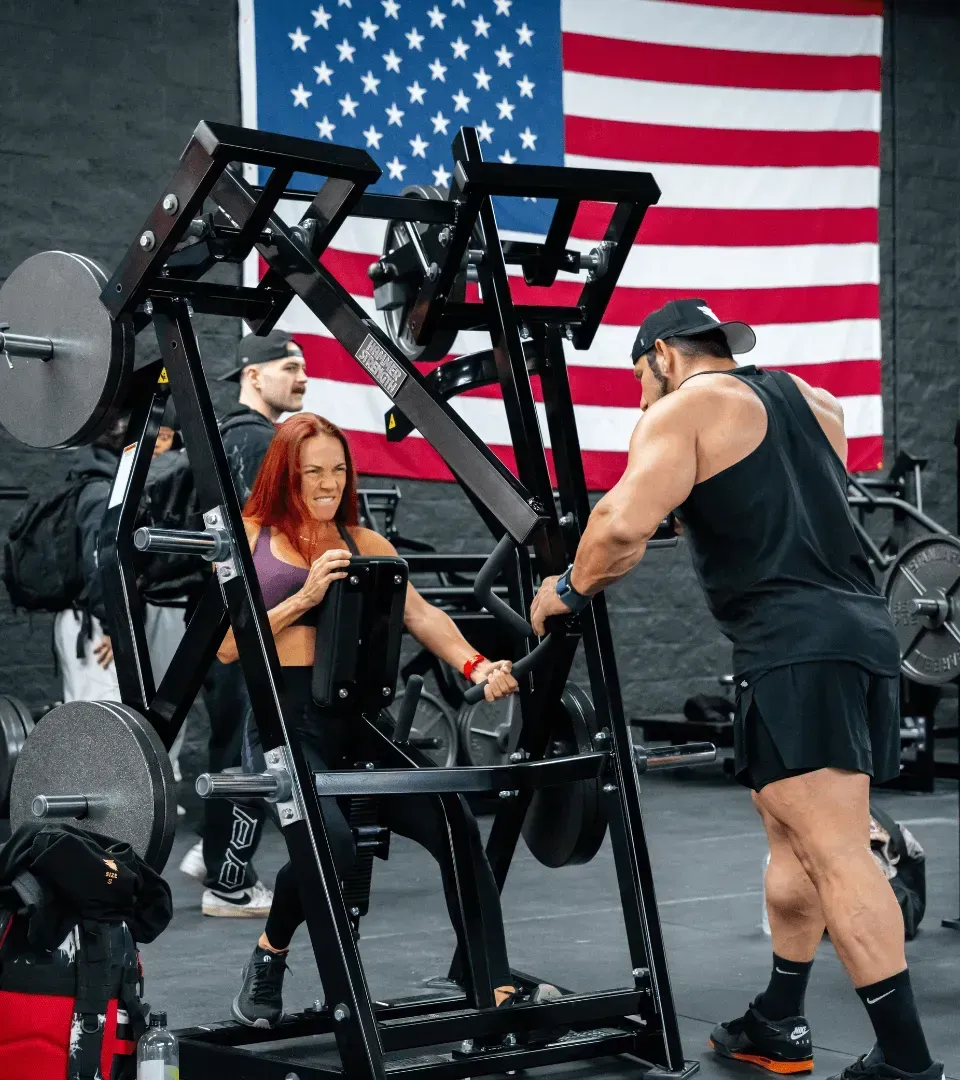 Two people using a strength machine in a gym, with an American flag hanging in the background.
