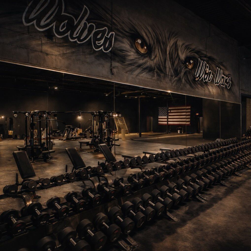 Dimly lit gym interior with rows of dumbbells, benches, and workout machines beneath a wolf mural.