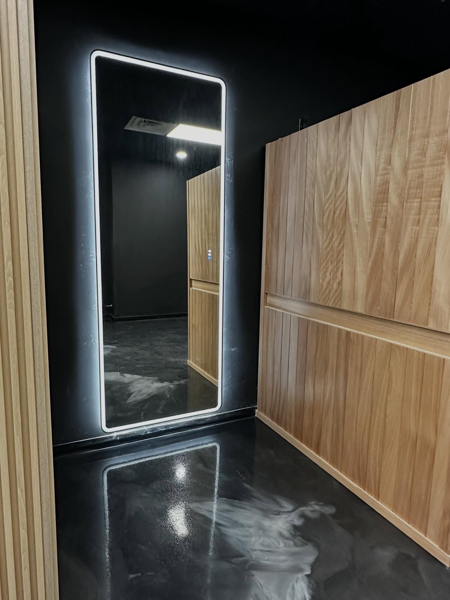 Modern hallway with a glowing full-length mirror, black walls, wood paneling, and a glossy floor.