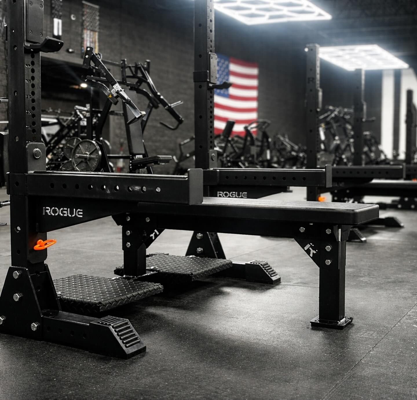 Black gym bench in a weight room with barbells, racks, and an American flag on the wall