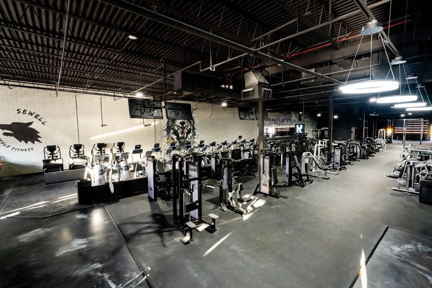 Large indoor gym with rows of weight machines, black flooring, and bright overhead lights