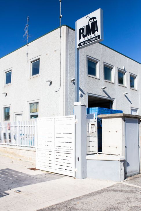 Edificio bianco a due piani con un’insegna sul tetto e un ingresso recintato, su una strada soleggiata.