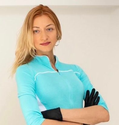 A person with blonde hair wearing a blue zip-up shirt and black gloves, posing with arms crossed against a white wall.