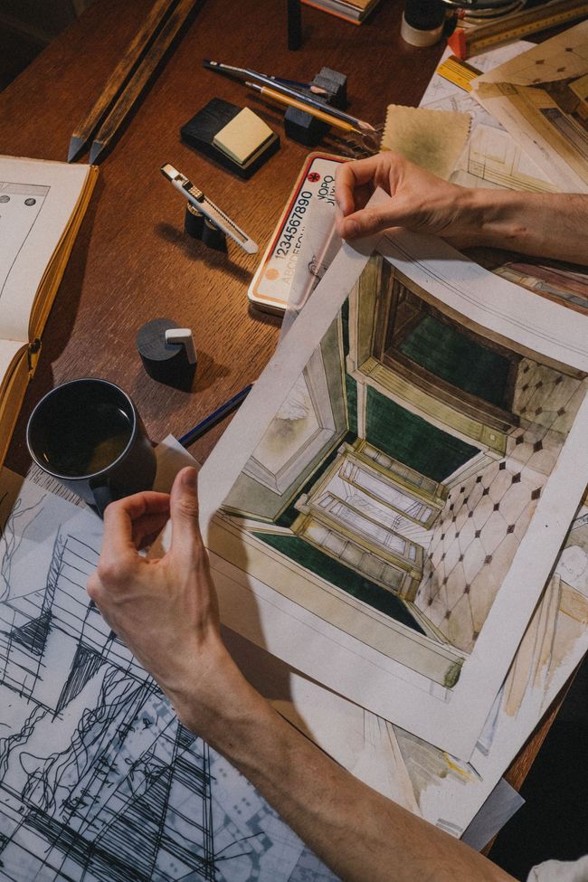 Hands sketching a small architectural model on a cluttered wooden desk with tools and paper