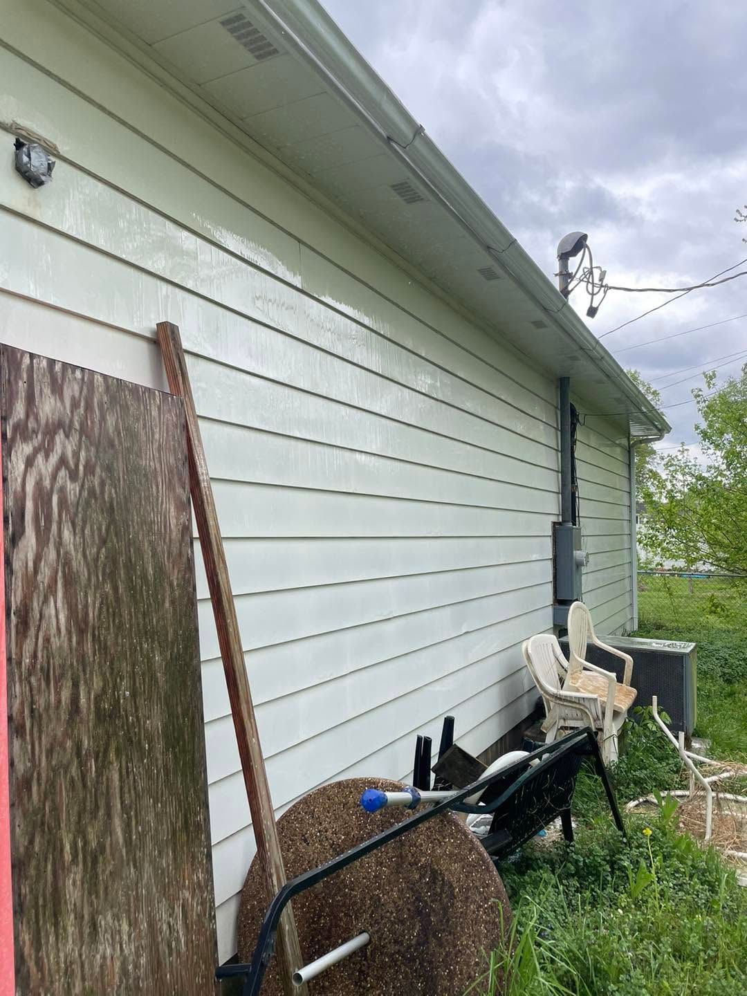 Side of a white house with stacked outdoor items, a chair, and yard tools under a cloudy sky