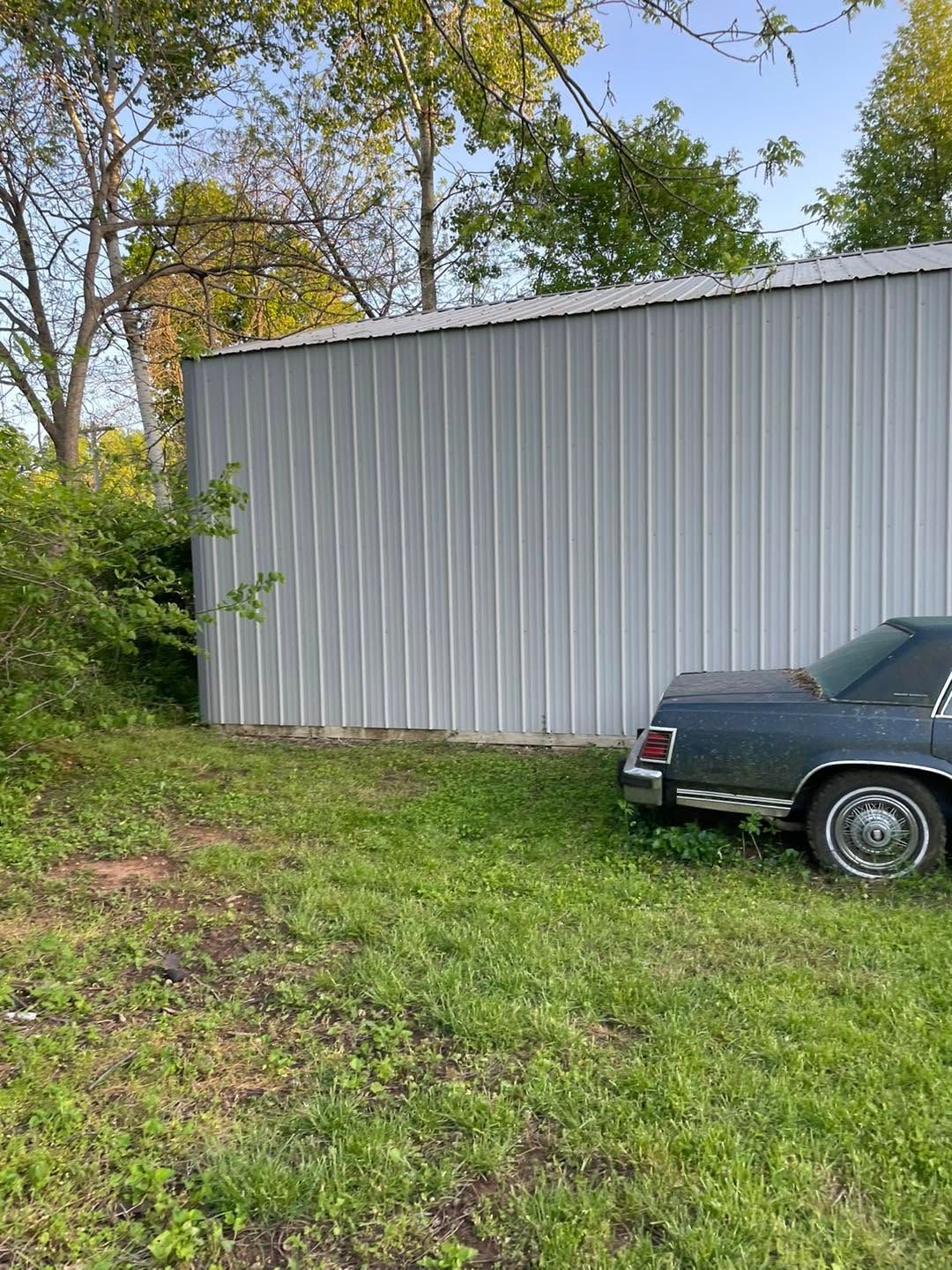 White metal building beside a grassy yard with trees and part of a dark car parked at right