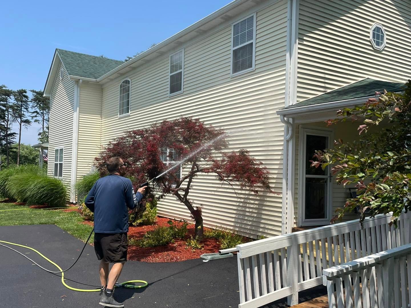 Person walking on a driveway beside a cream-sided house and red-leafed shrub, near a white fence