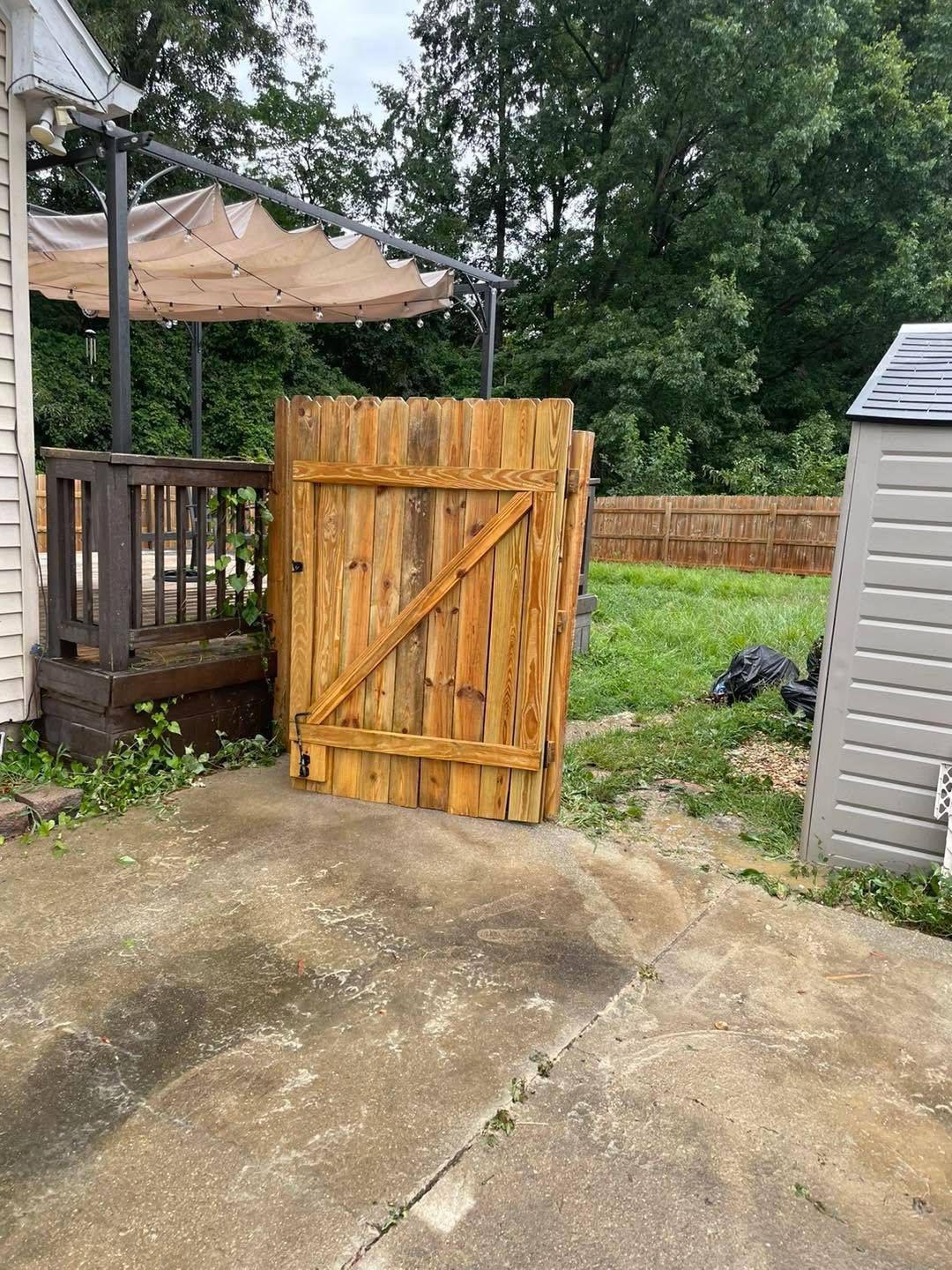 Wooden gate leaning open between a porch and shed, with a concrete patio and grassy yard beyond.