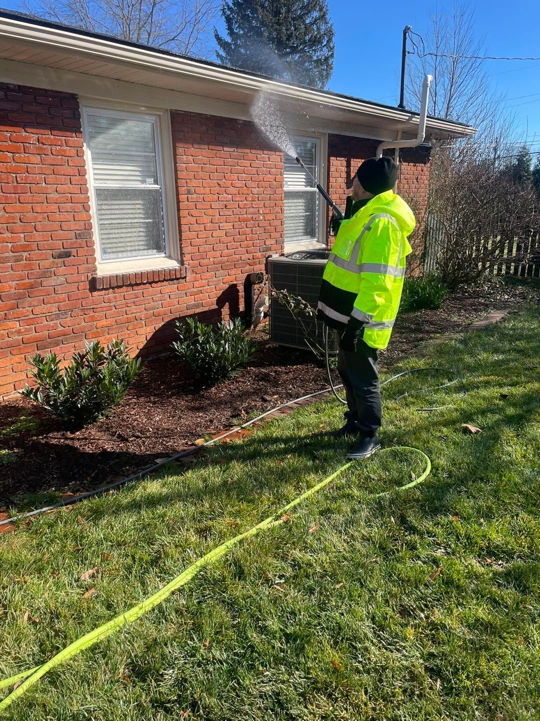 Worker in neon jacket standing by a brick house, using a hose in a sunny yard.