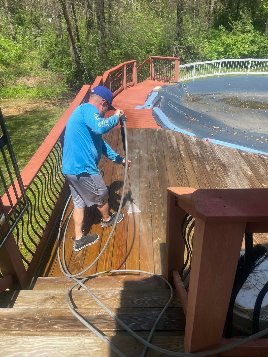 Worker pressure-washing a wooden deck beside an above-ground pool on a sunny day