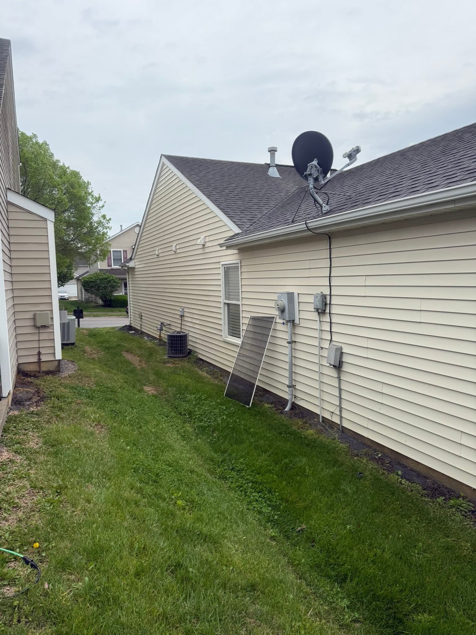 Side view of a beige house with roof damage, showing warped siding and a grassy narrow side yard.