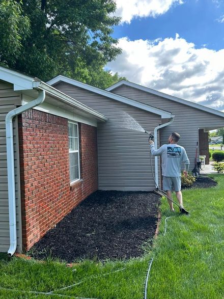 Worker spraying house exterior siding with a hose beside brick and gray walls, under a blue sky