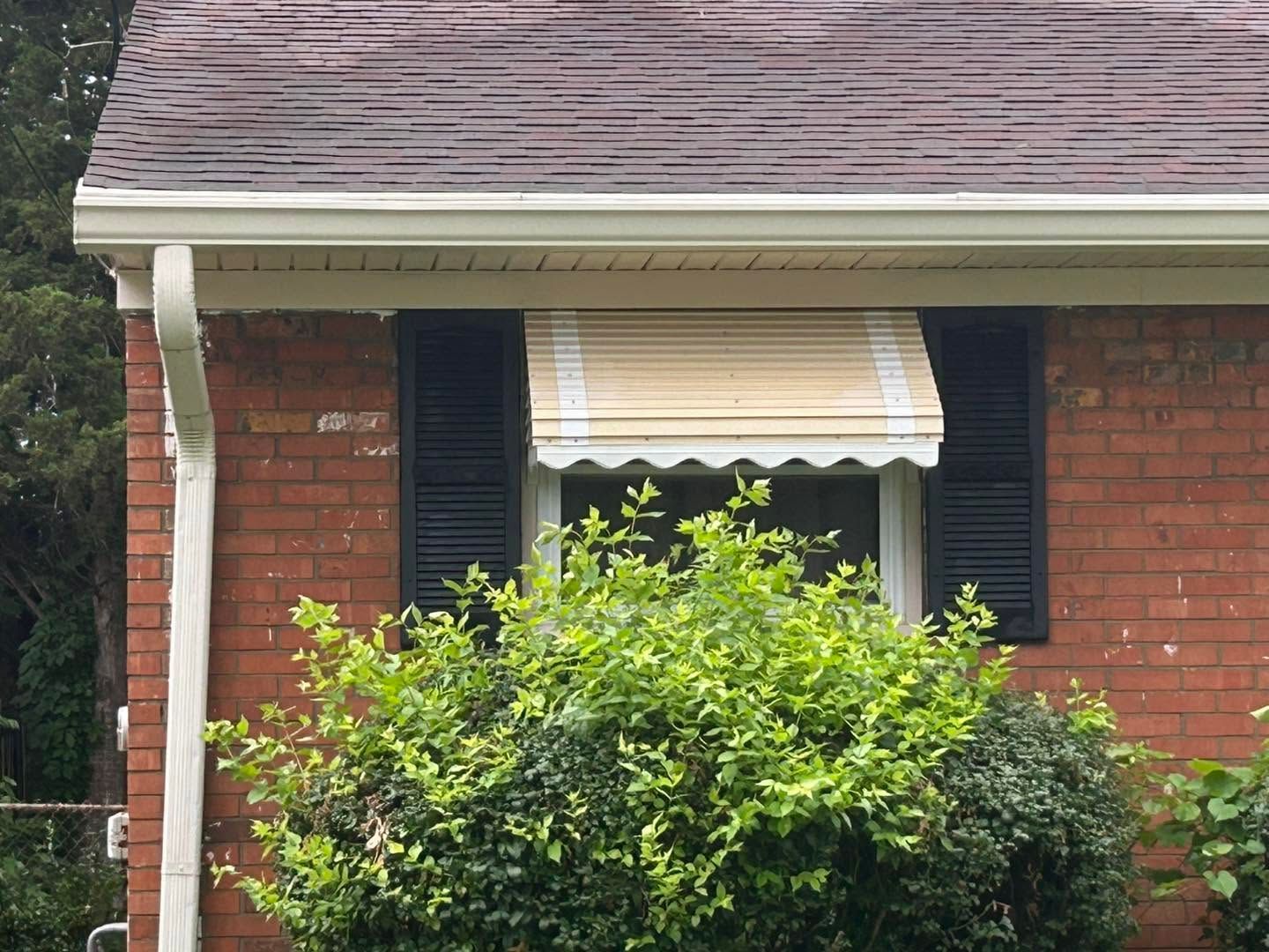 Brick house front with black shutters, striped awning, and dense green bushes under the window